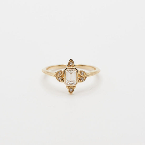 emerald cut diamond ring with milgrain and diamond accents on each side in yellow gold on white background