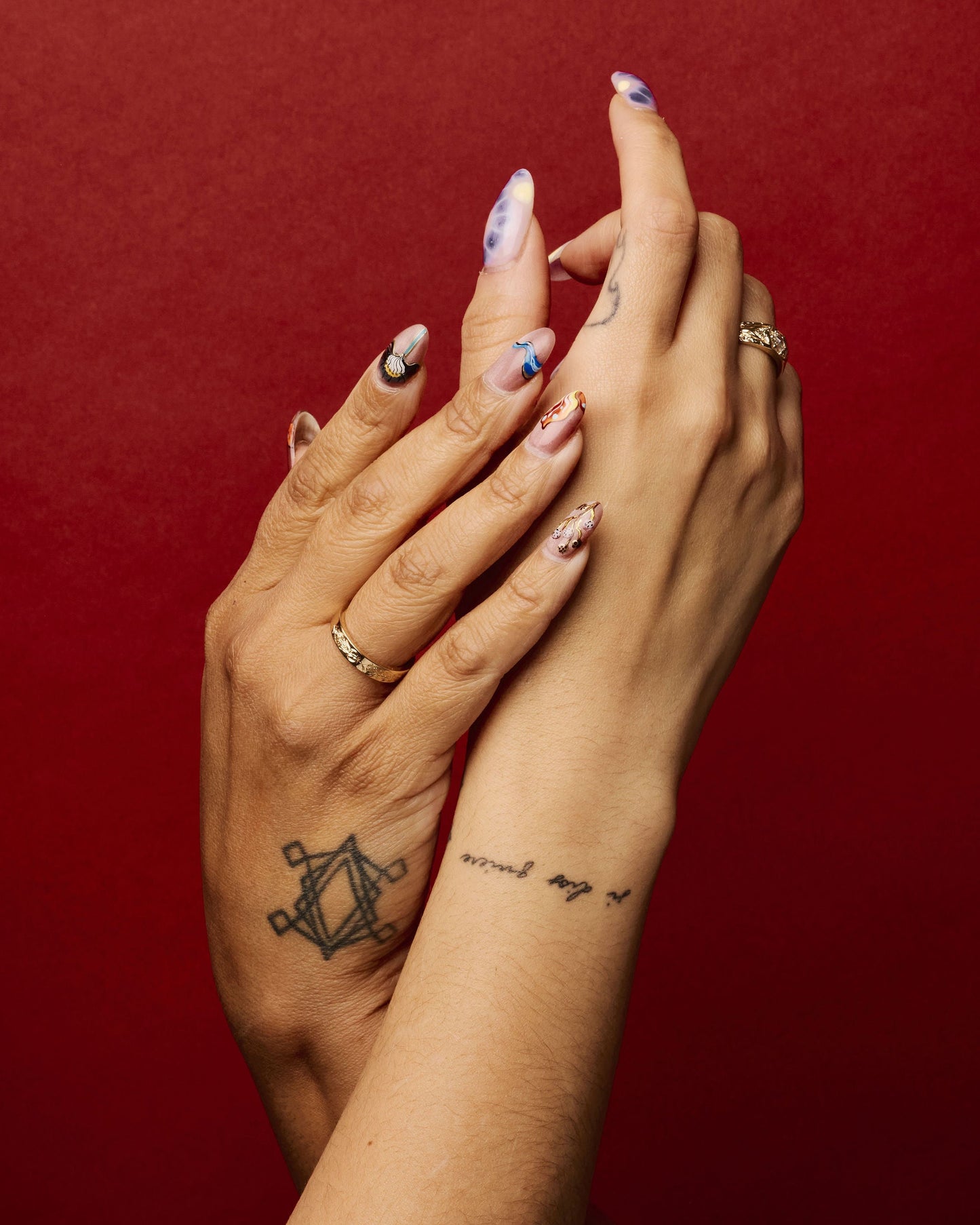 two hands intertwined on a red background, one wearing the element 5 ring on ring finger, the other wearing another engraved gold band on their ring finger.
